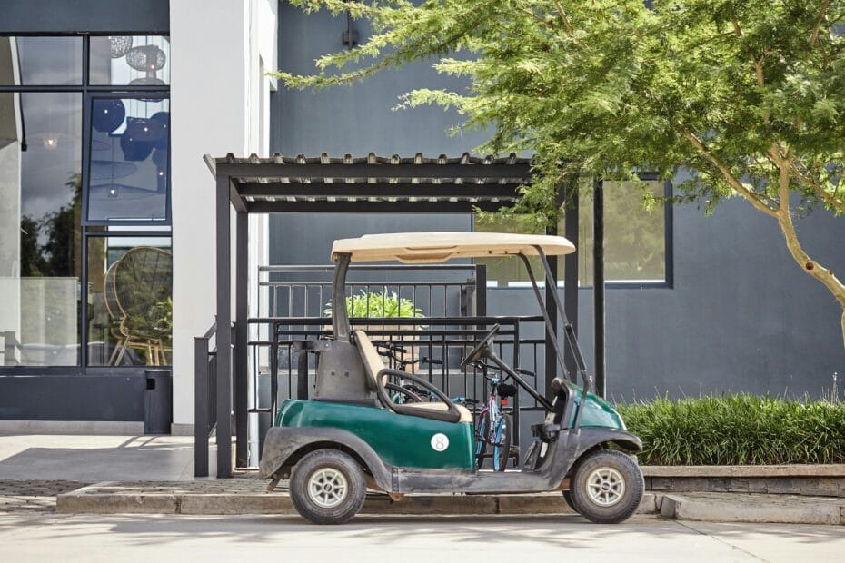 A green electric golf trolley parked at a stylish, modern resort entrance, surrounded by greenery.