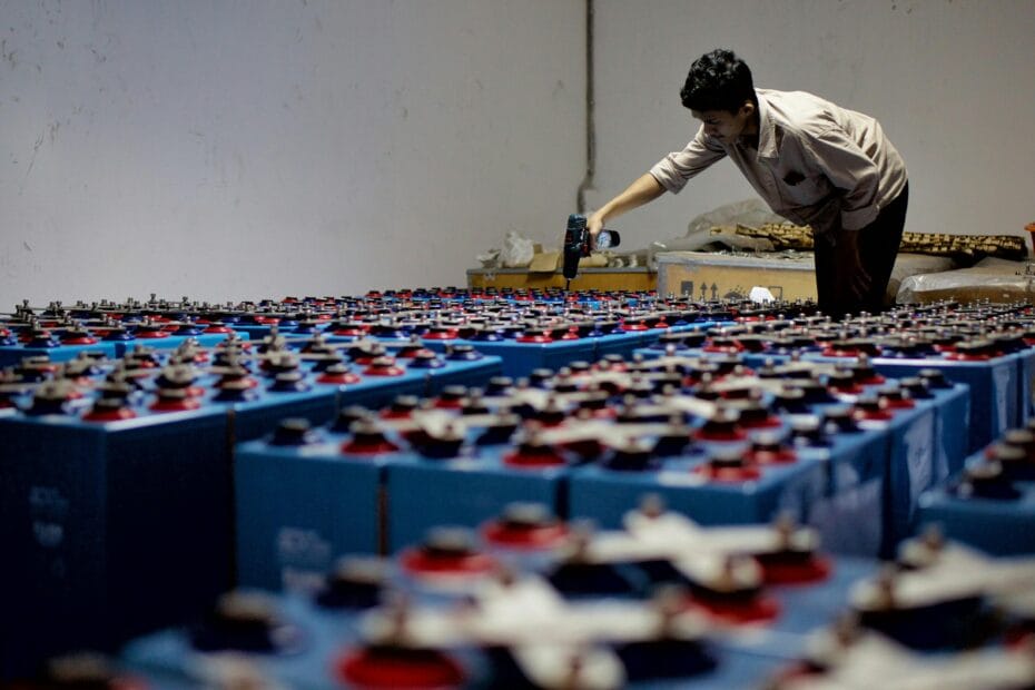 A worker checking many industrial batteries inside a facility. Indoor, industrial setting.