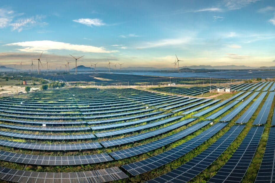 Ausgedehnter Solarpark mit Windturbinen im Hintergrund unter einem klaren blauen Himmel.