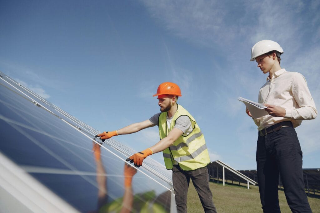 From below of bearded electrical technician in yellow vest and hardhat demonstrating solar panels to young male inspector in formal wear